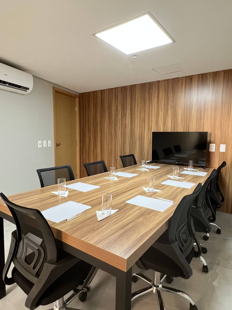 Sala de conferências com mesa longa de madeira, cadeiras pretas, TV e parede com painéis de madeira.