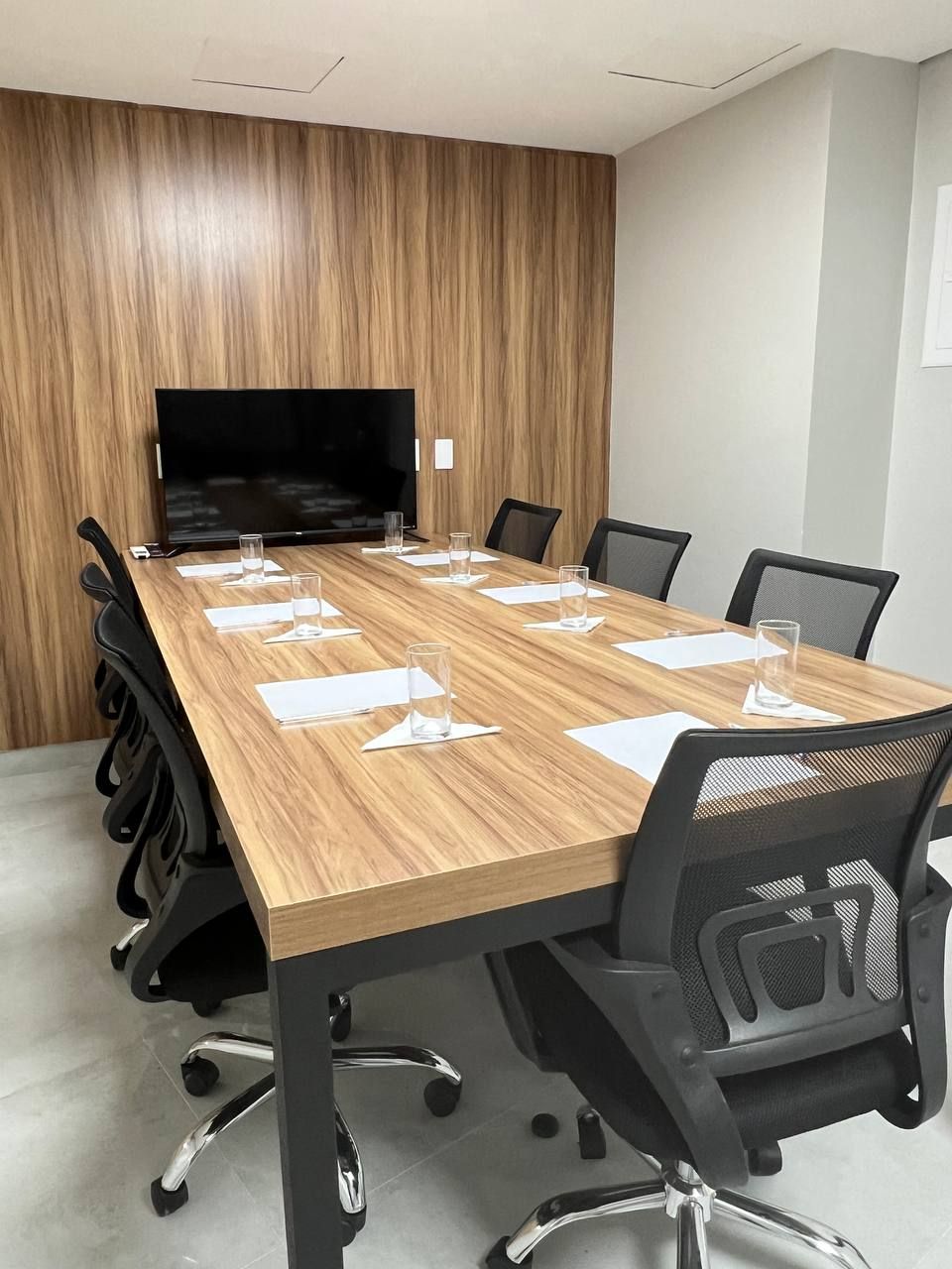 Sala de conferências com uma grande mesa de madeira, cadeiras e uma TV em uma parede de painéis de madeira.