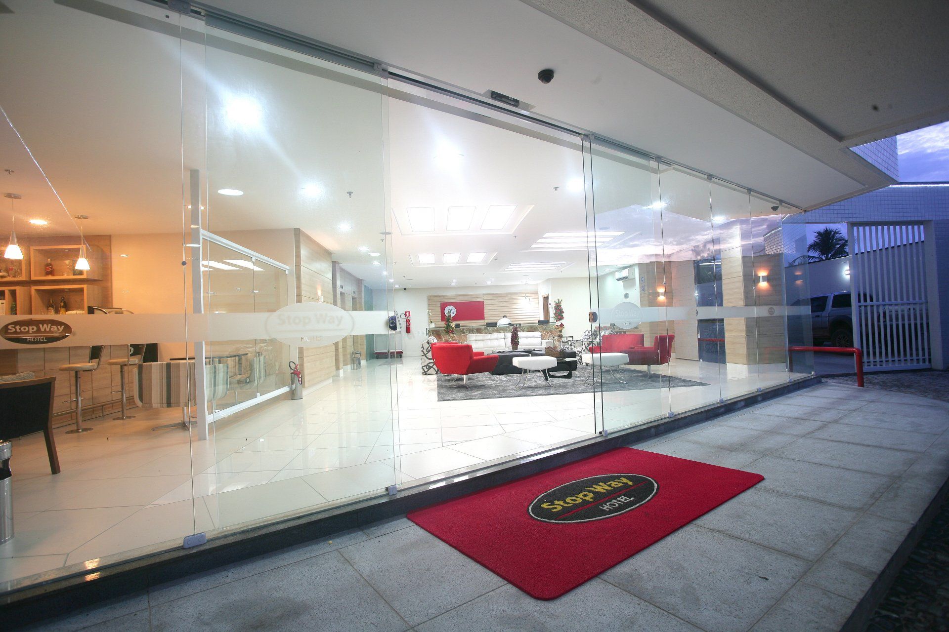 Entrada para um saguão moderno com capacho vermelho. Grandes portas de vidro revelam um interior iluminado e uma área de estar.