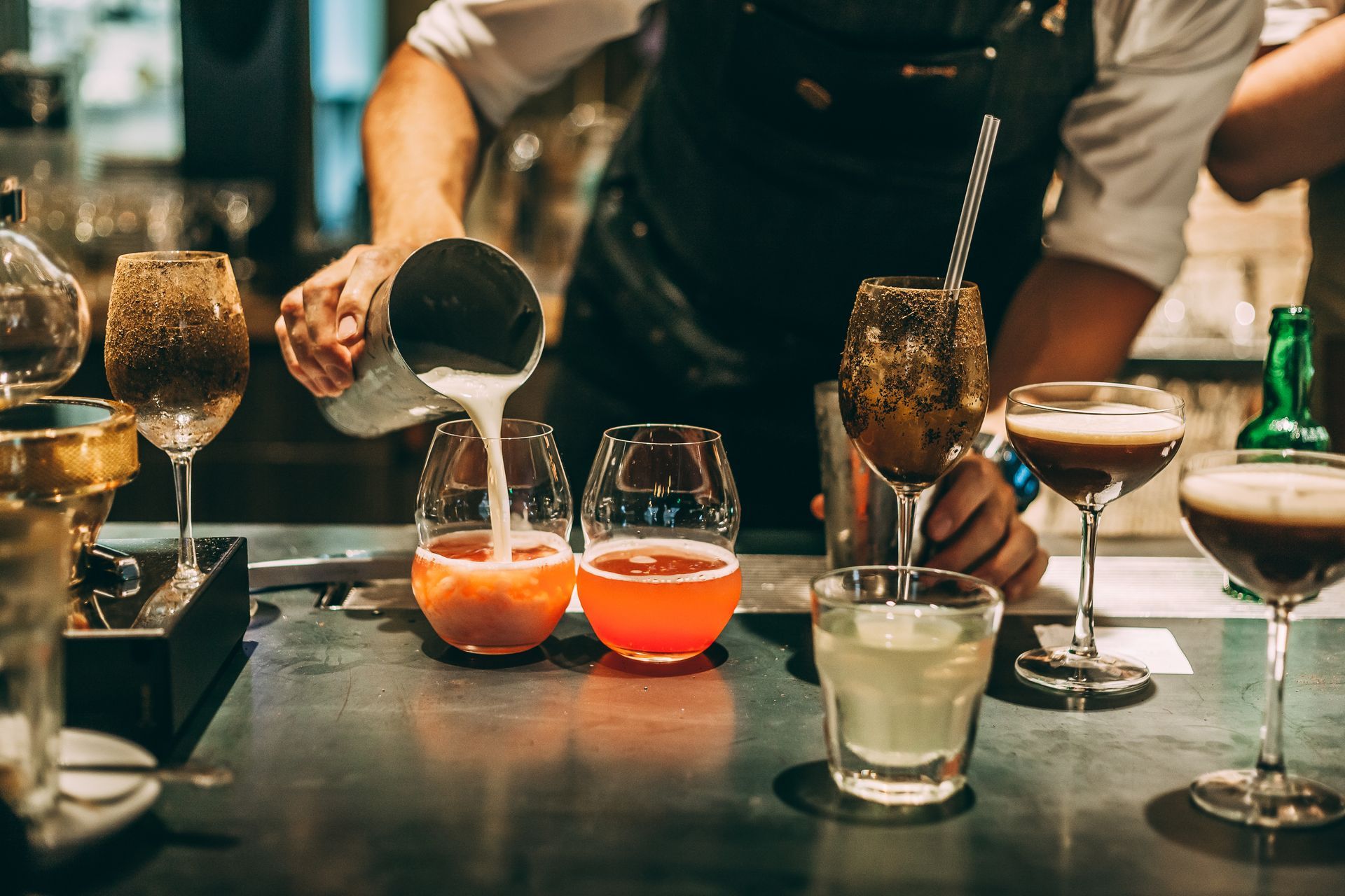 Bartender pouring a white drink into glasses with colorful cocktails on a bar counter