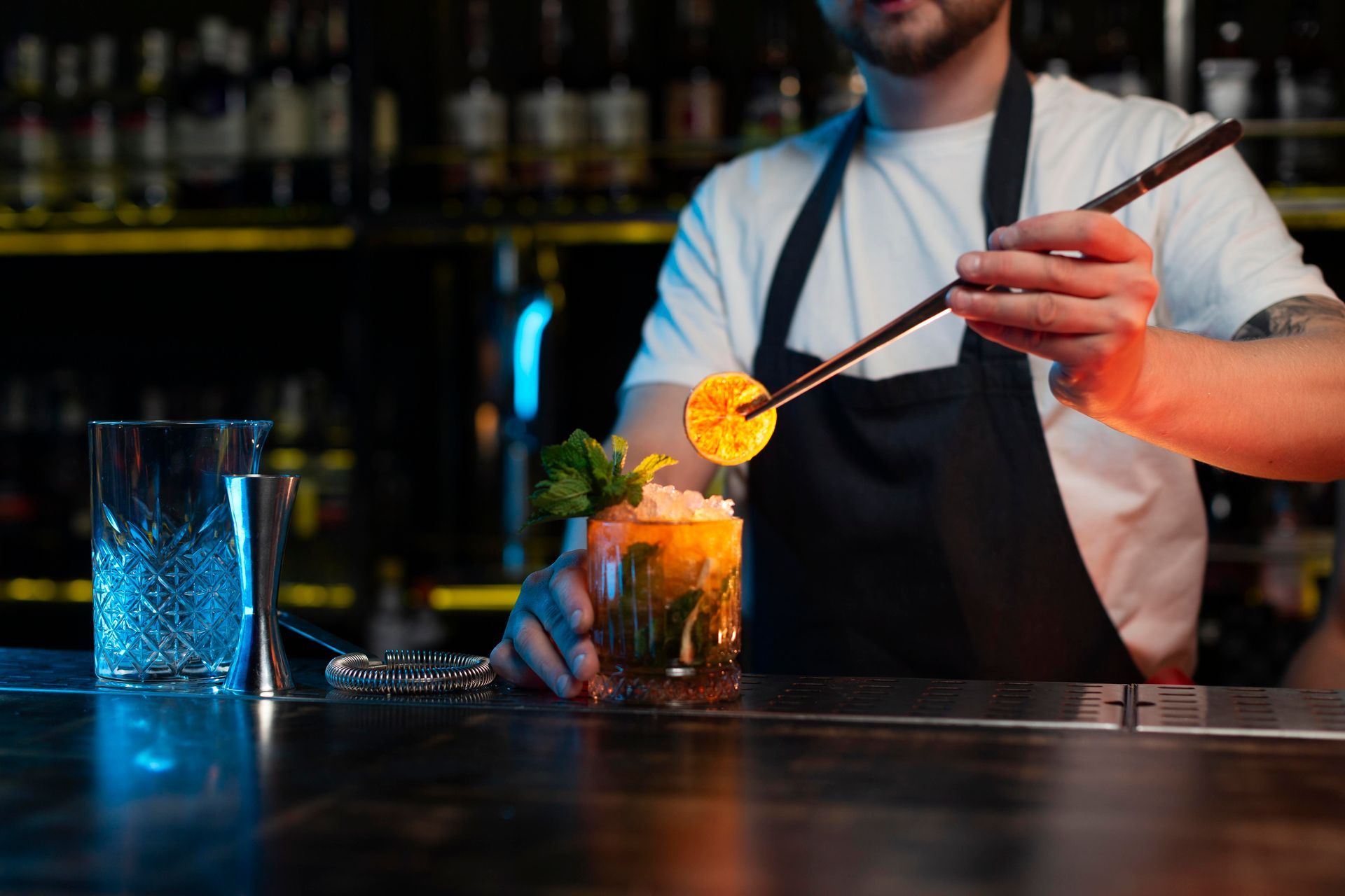 Bartender garnishes a cocktail with orange slice. Bar setting, low light.