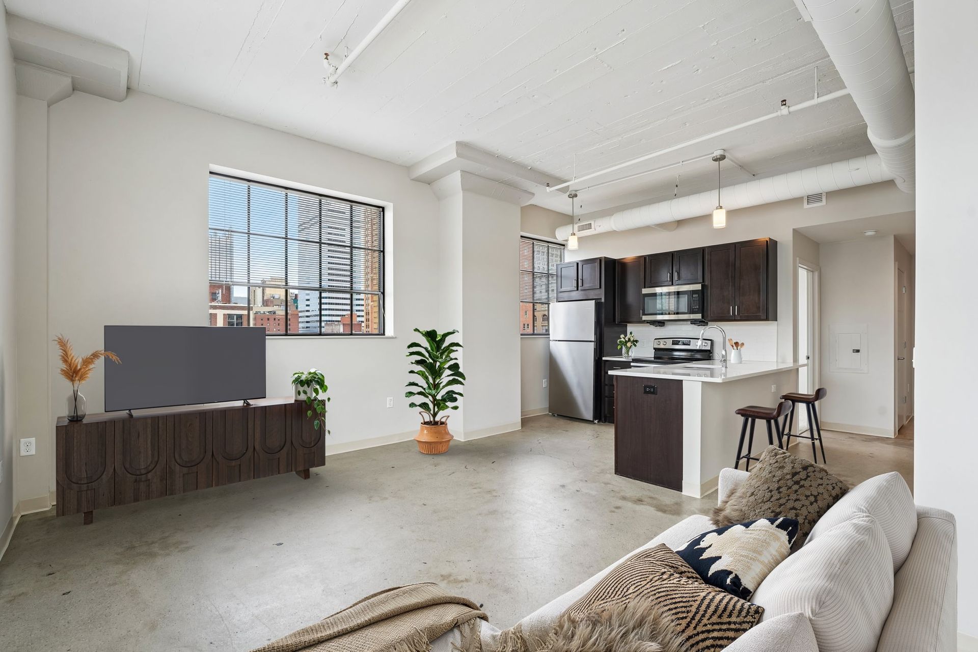 Bright open loft living room with sectional sofa, TV console, kitchen island, and large windows at Terminal 21, a premier apartment community in Downtown Pittsburgh, Pennsylvania.