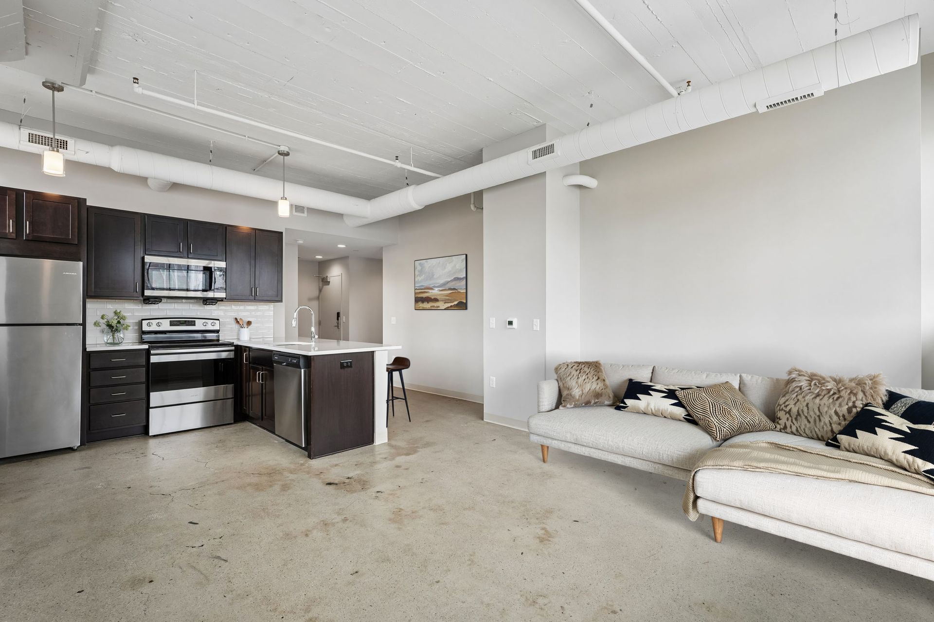 Bright open loft living room with kitchen, gray sofa, stainless appliances, and high ceilings at Terminal 21, a premier apartment community in Downtown Pittsburgh, Pennsylvania.