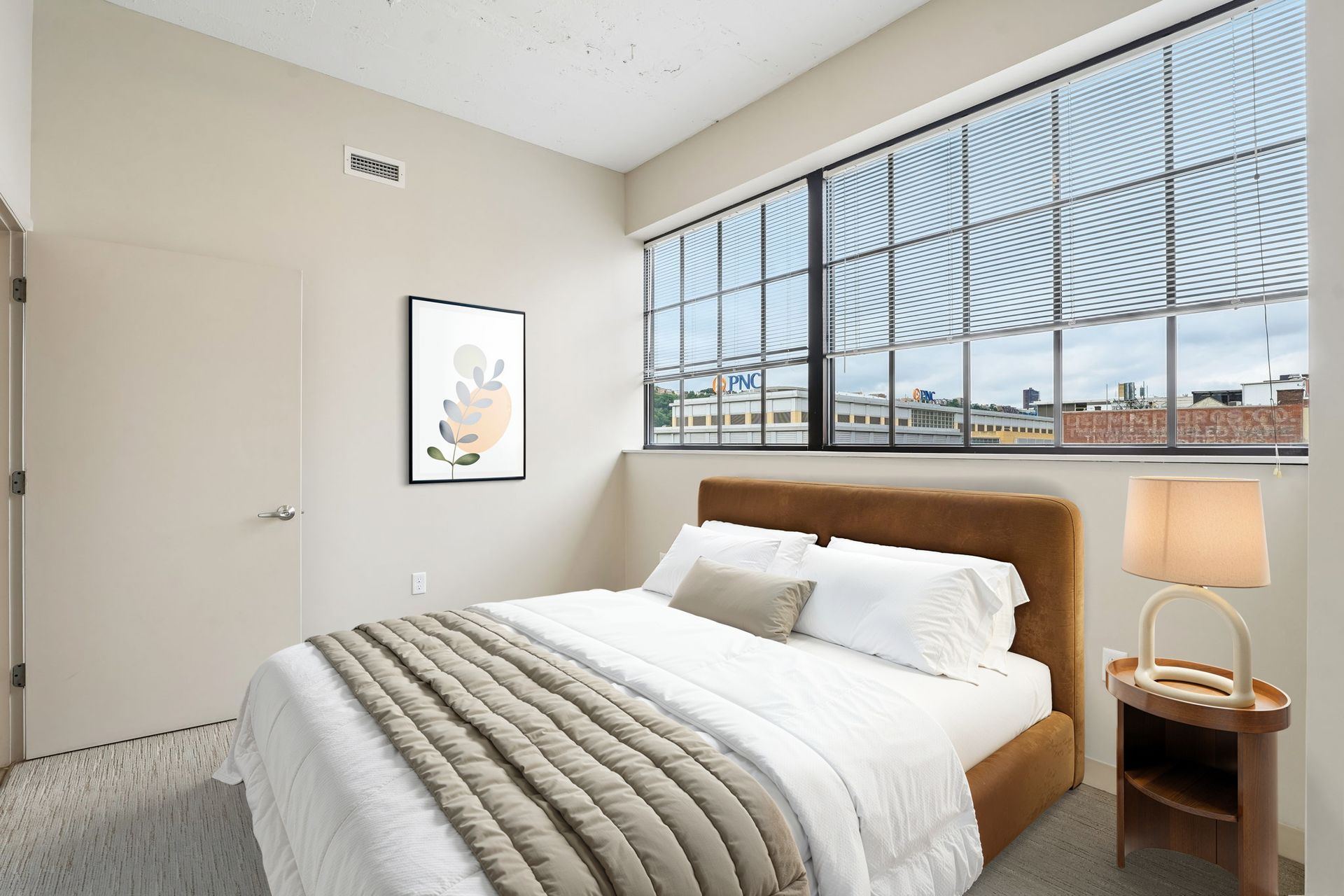 Bright bedroom with a bed, nightstand lamp, framed art, and large windows with city view at Terminal 21, a premier apartment community in Downtown Pittsburgh, Pennsylvania.