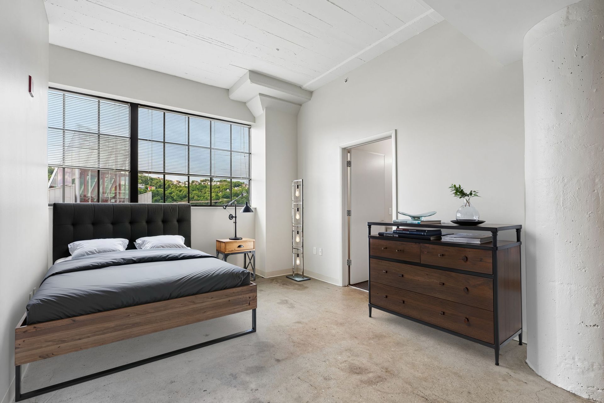 Bright studio apartment with black bed, wooden dresser, and large window blinds at Terminal 21, a premier apartment community in Downtown Pittsburgh, Pennsylvania.
