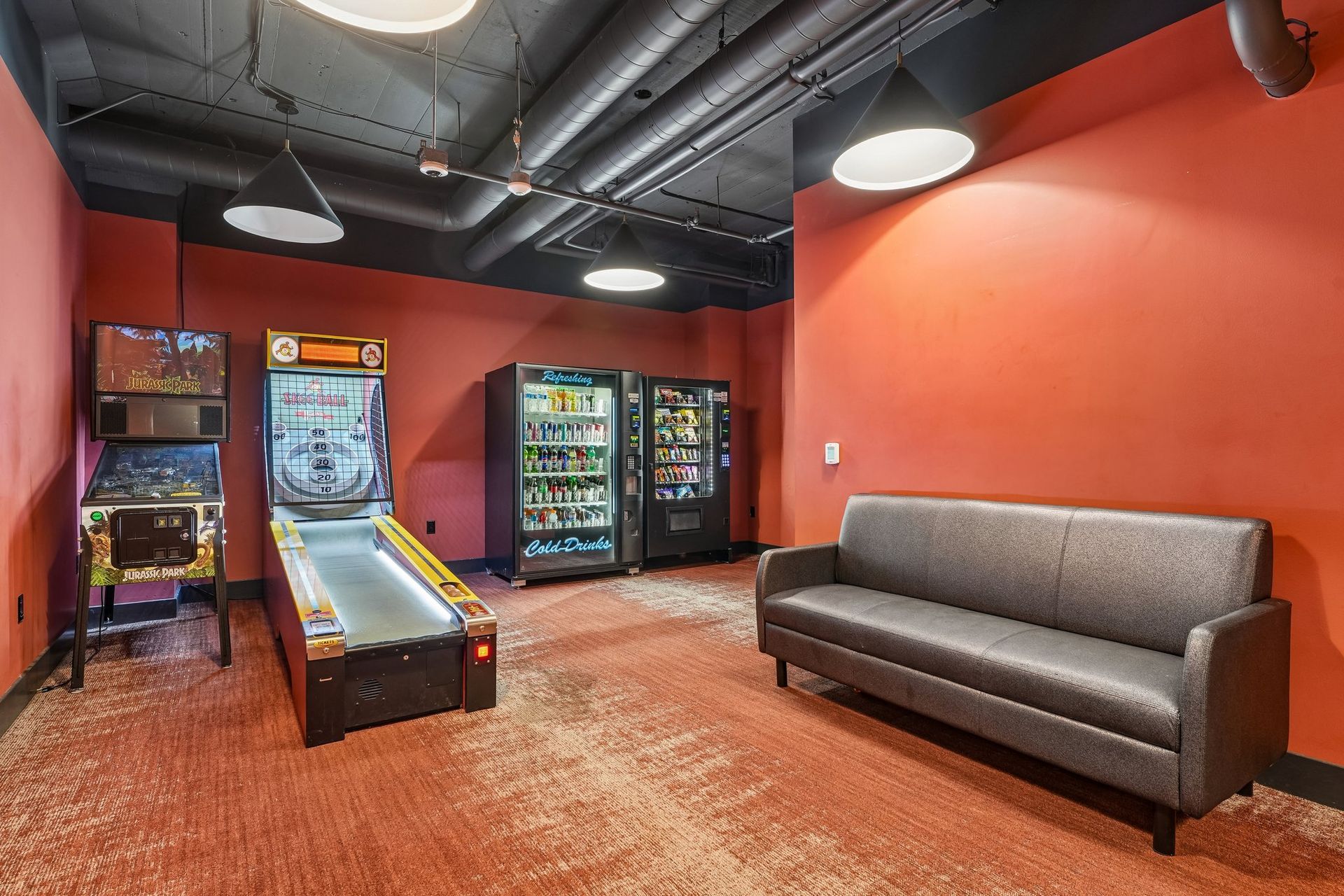 Red lounge with arcade games, vending machines, and a gray couch under industrial lighting at Terminal 21, a premier apartment community in Downtown Pittsburgh, Pennsylvania.