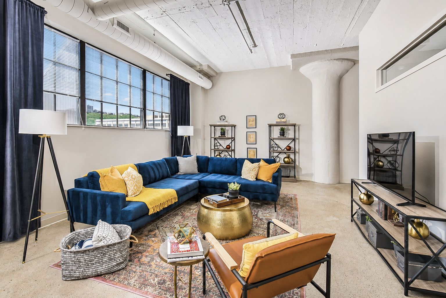 Bright living room with blue sectional, patterned rug, concrete floors, and large windows near a TV console at Terminal 21, a premier apartment community in Downtown Pittsburgh, Pennsylvania.