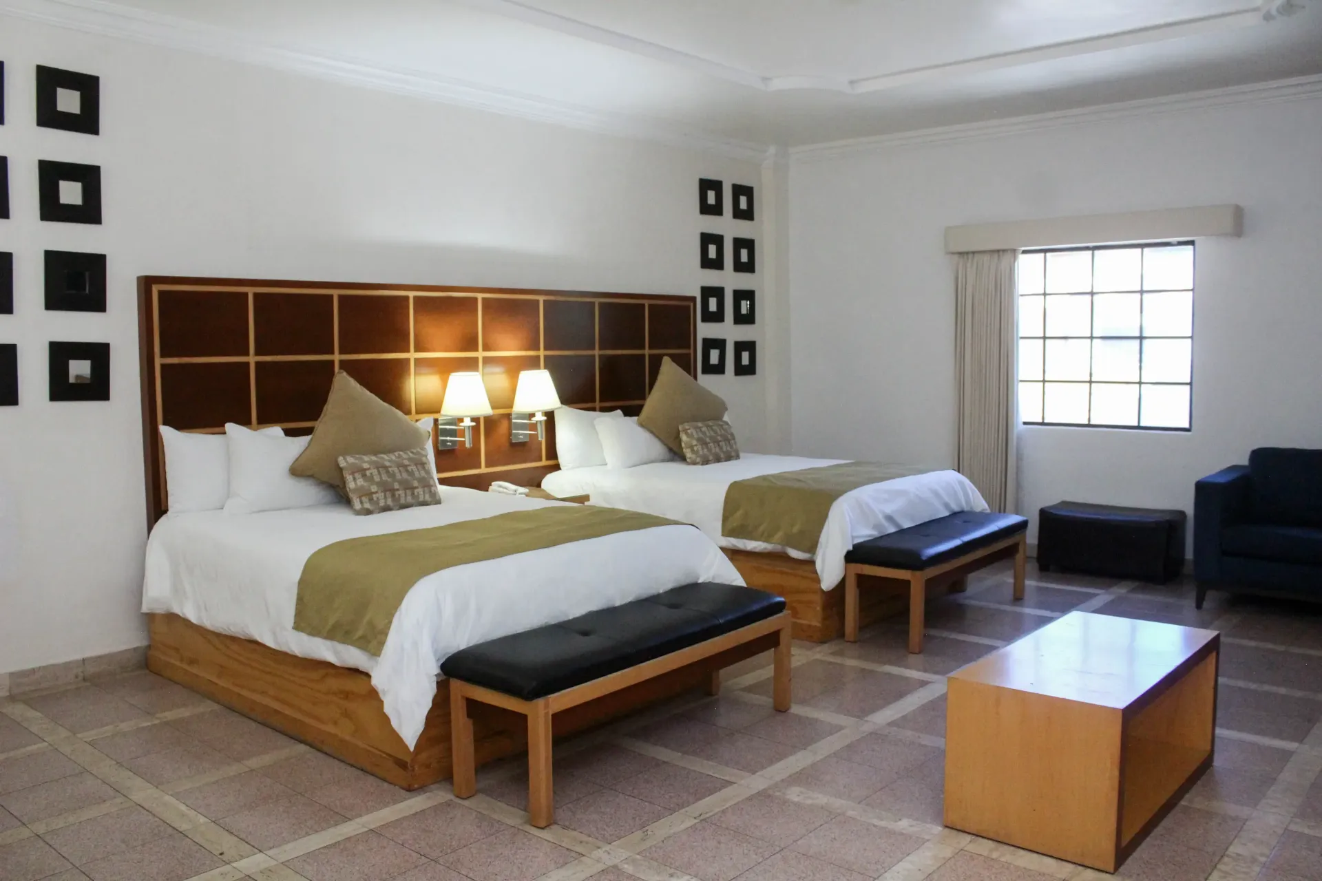 Habitación de hotel con dos camas, ventana y zona de estar. Tonos neutros, bien iluminada.