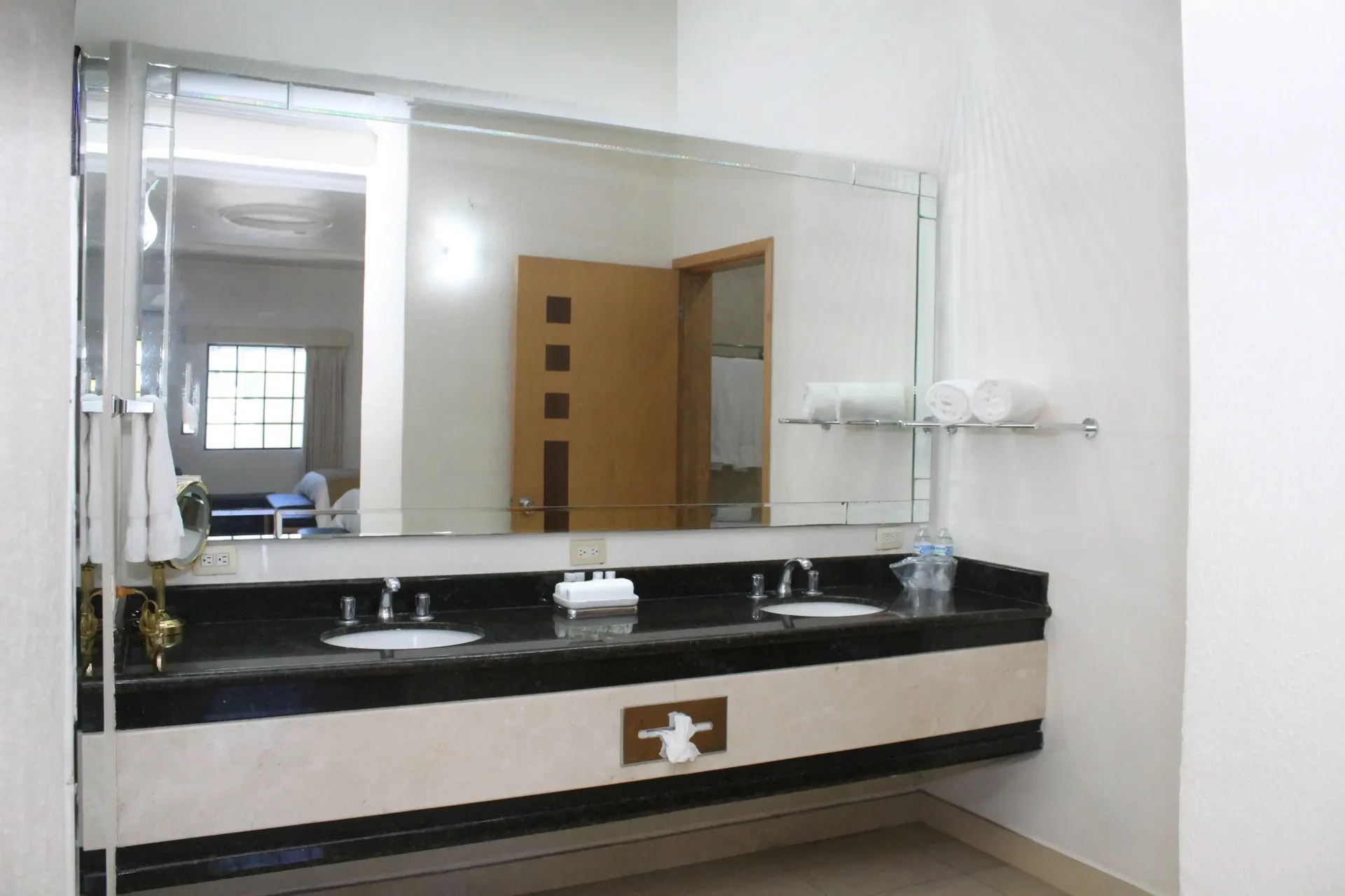 Baño con lavabo doble, espejo grande y puerta abierta. Encimera negra, paredes claras y toallas.