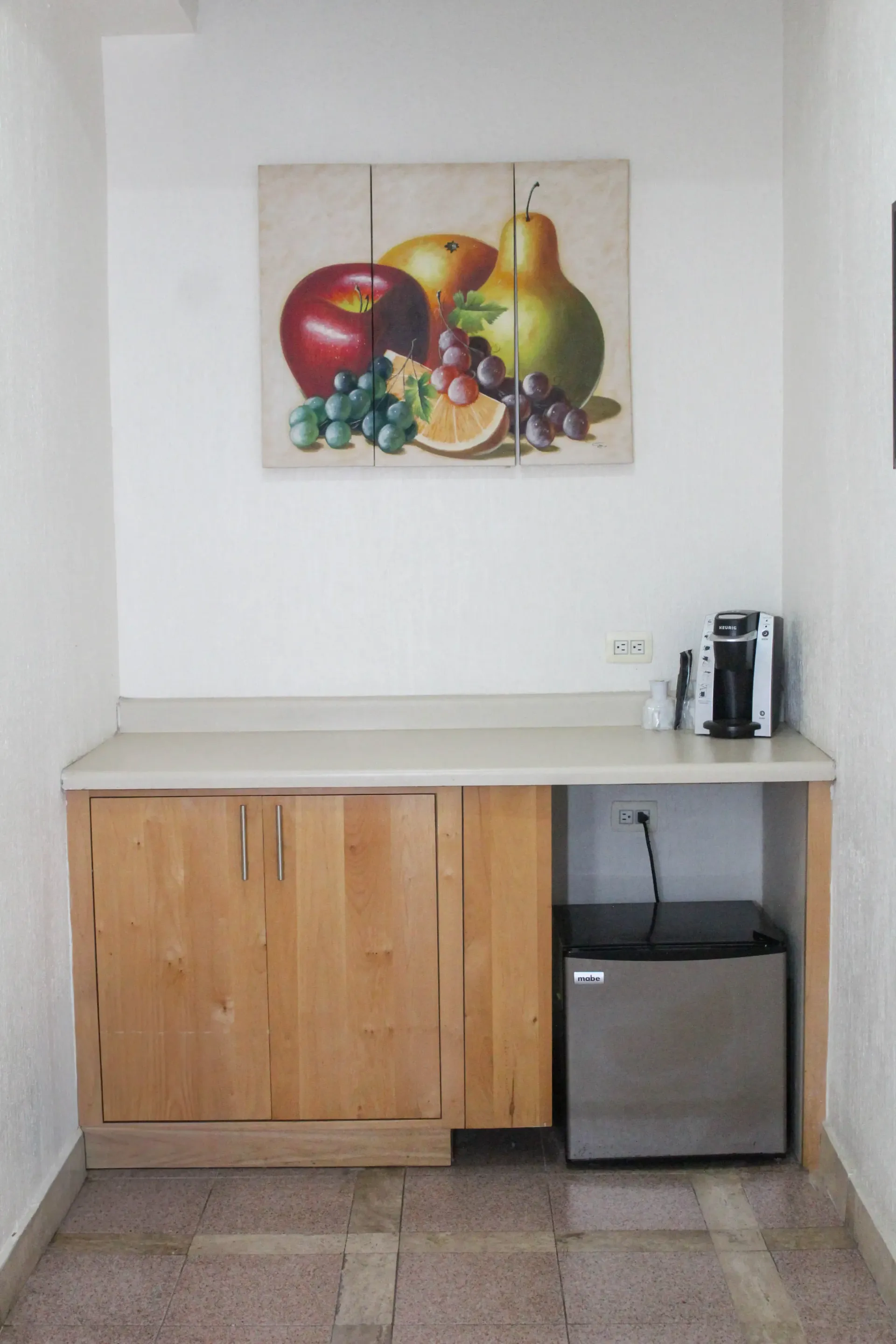 Armario empotrado con mininevera y encimera, cafetera y bodegón de frutas en la pared.