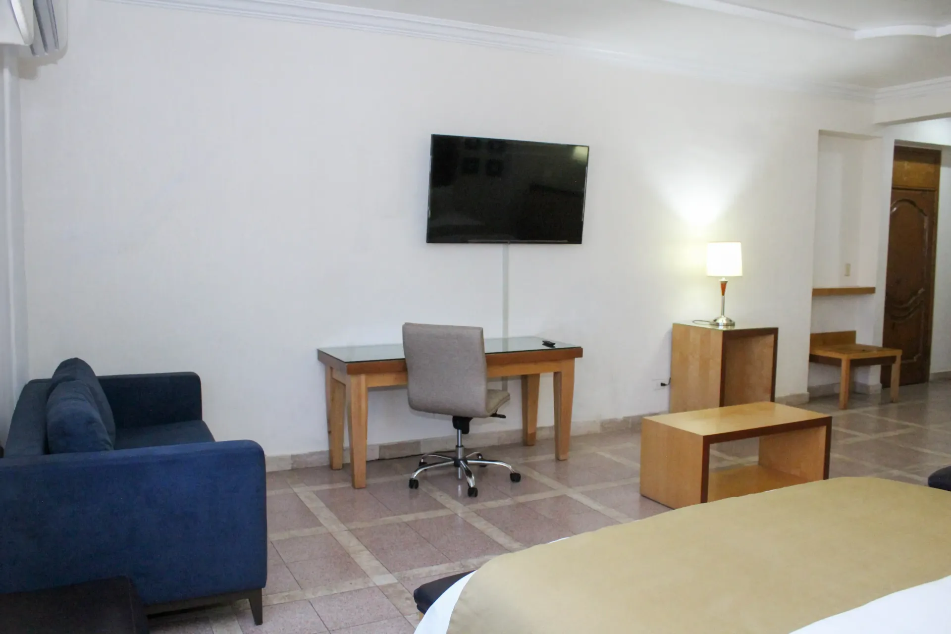 Sala de estar con sofá azul, escritorio, televisor y lámpara; suelo de baldosas.