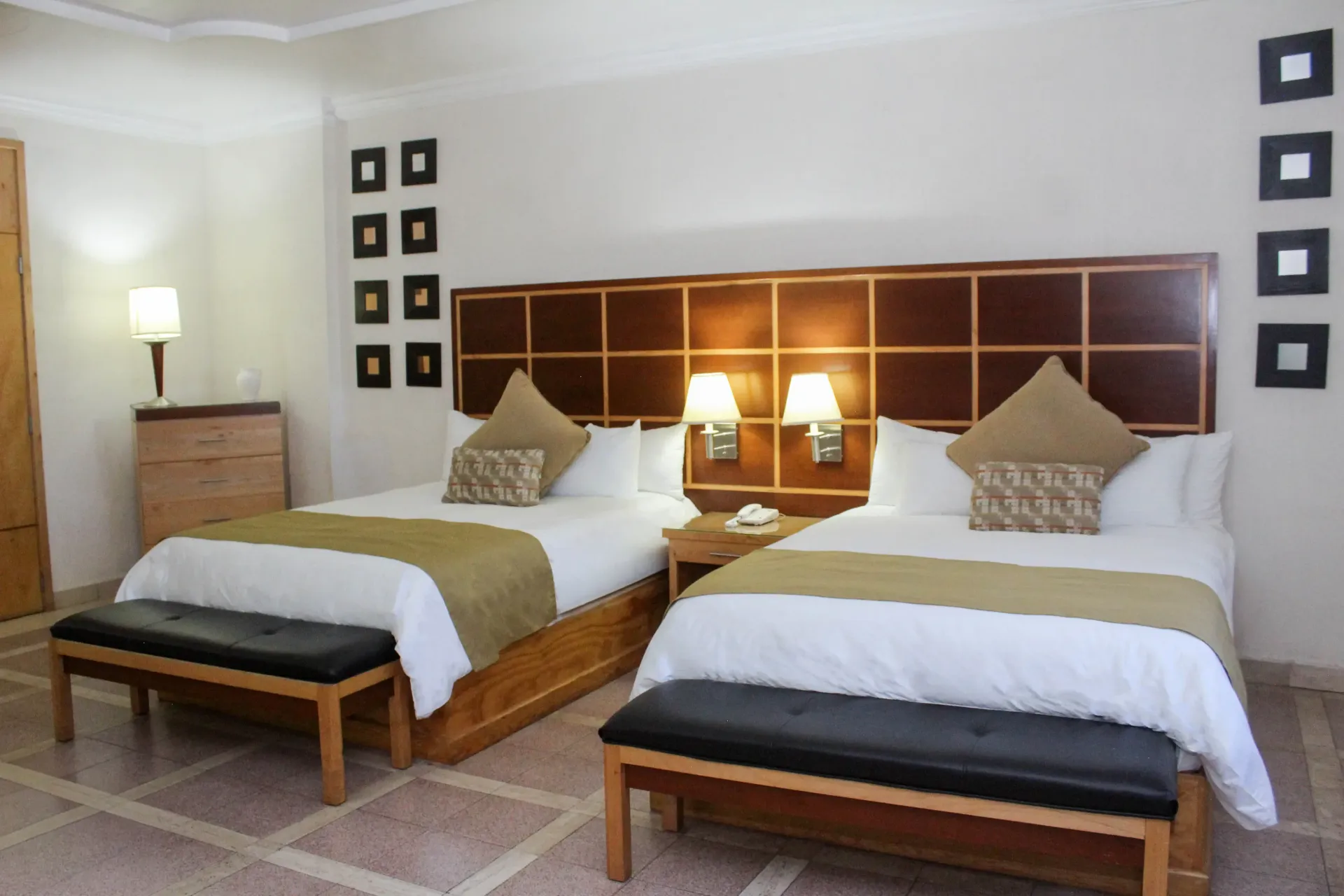Habitación de hotel con dos camas, cabecero marrón, mesitas de noche, lámparas y un banco.