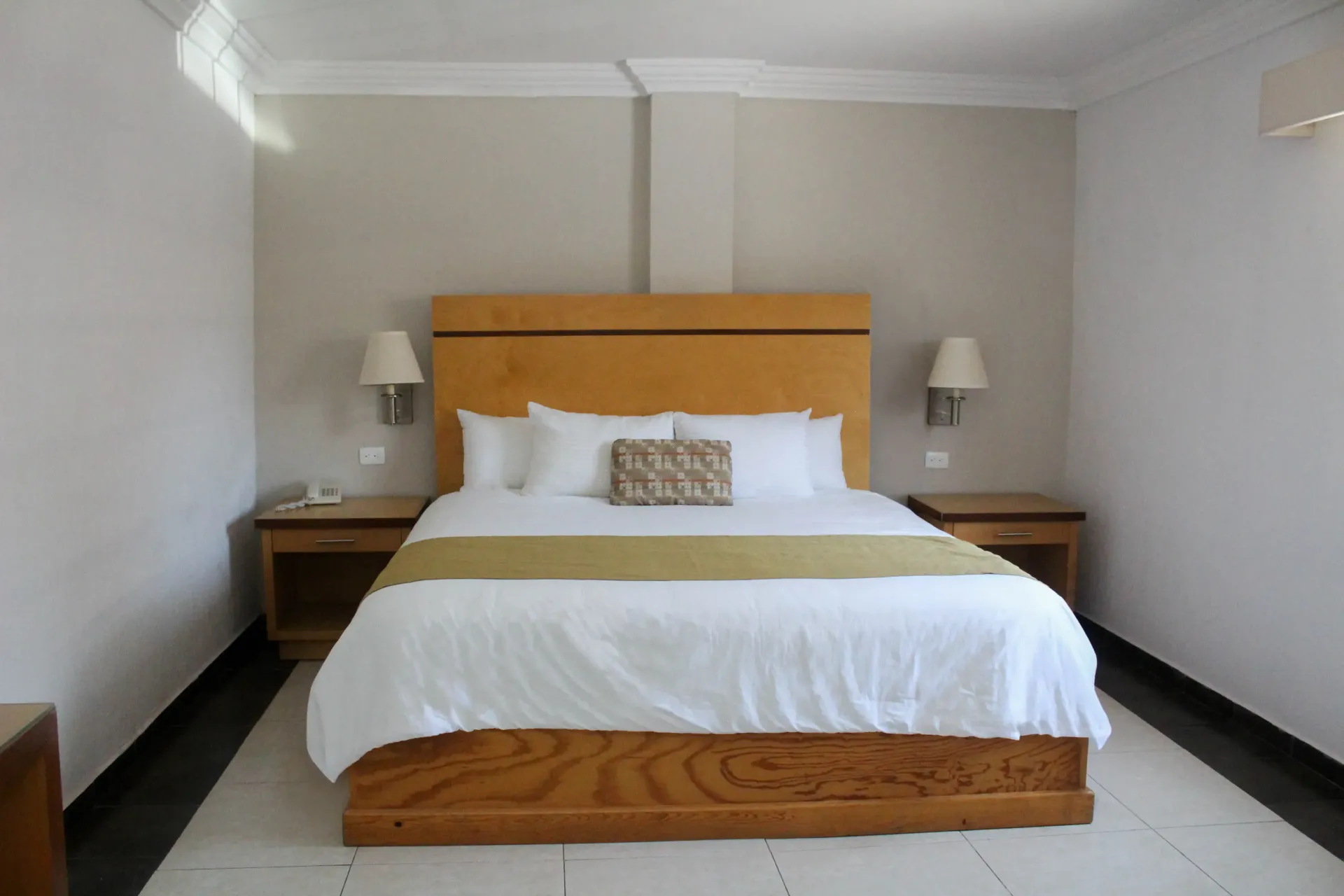 Habitación de hotel con una gran cama de madera, dos mesitas de noche y apliques de pared. Tonos beige y blancos.
