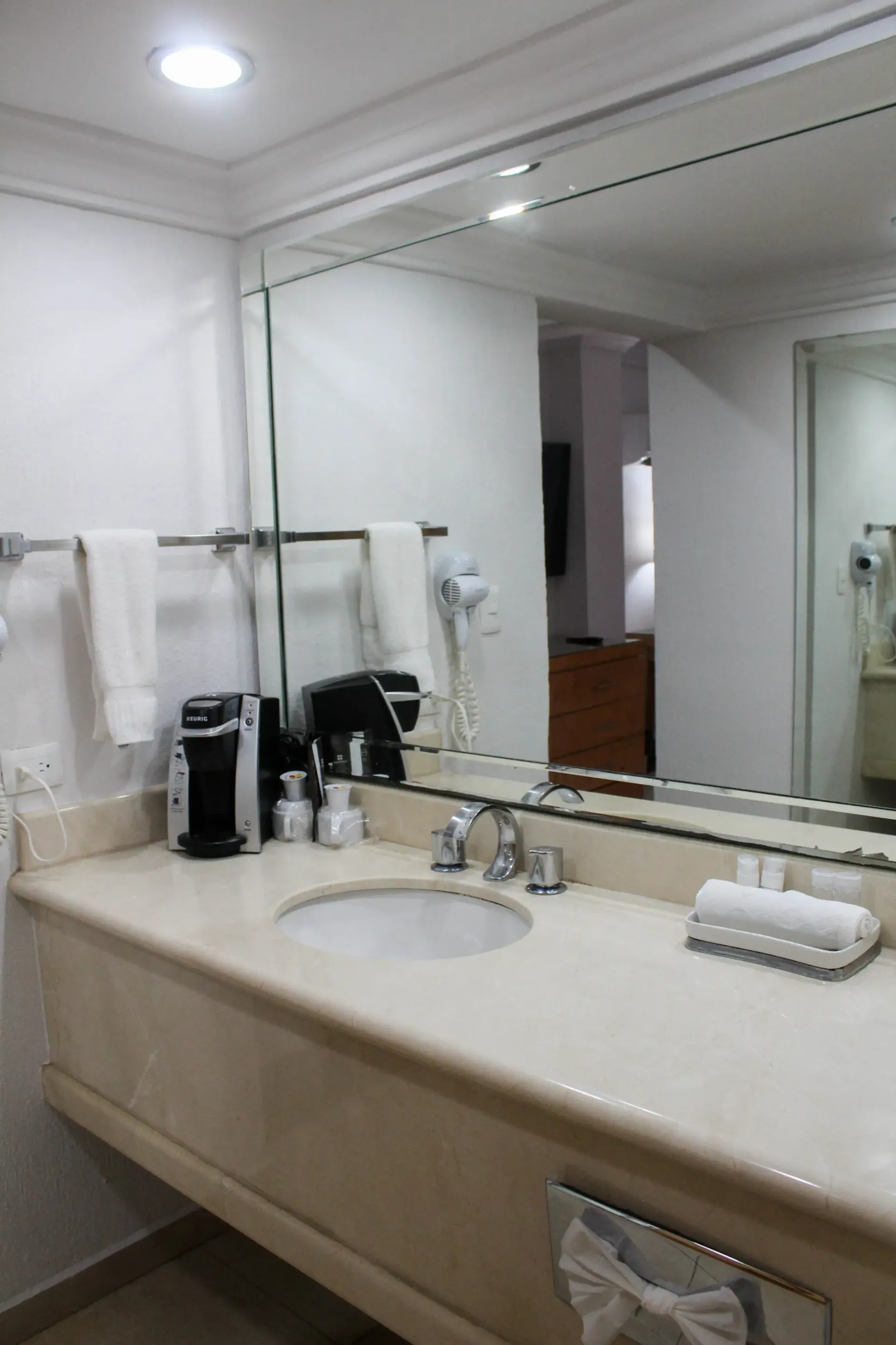 Baño con un gran espejo sobre una encimera de mármol beige claro con lavabo, artículos de aseo y toallas.