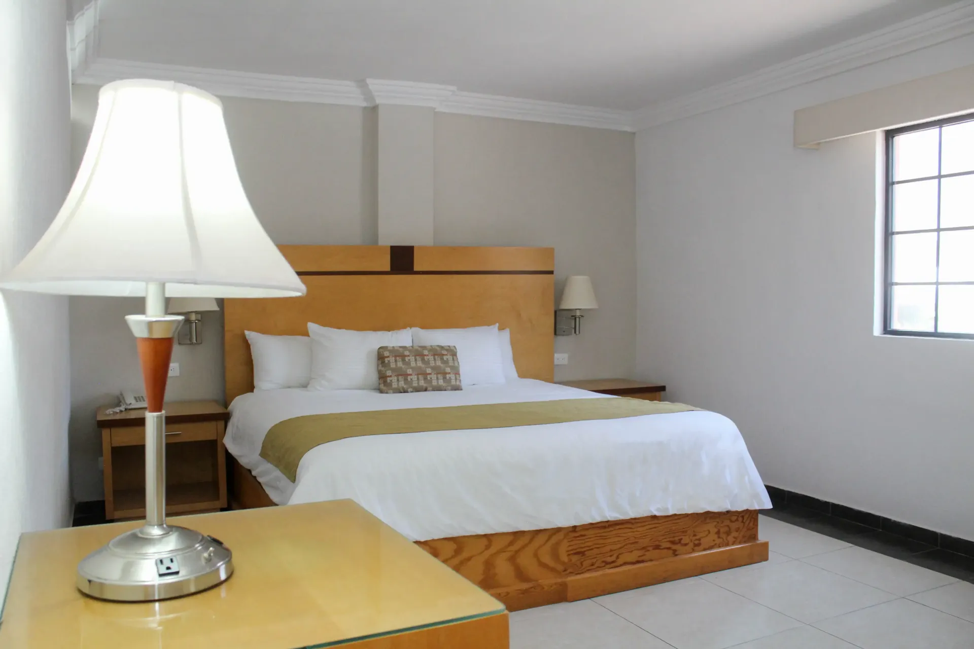Habitación de hotel con cama matrimonial, mesitas de noche y lámpara. Ventana a la derecha.