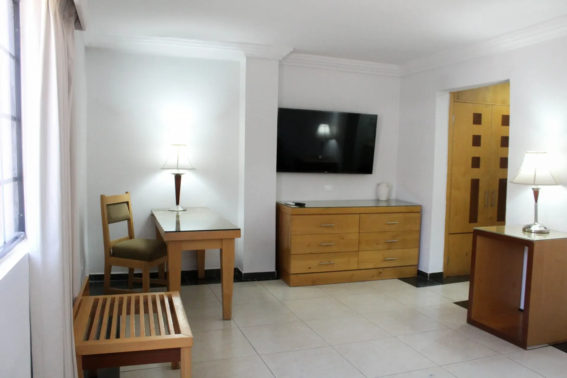 Una habitación de hotel con televisor, escritorio y muebles de madera. Paredes blancas, suelo de baldosas y puerta cerrada.