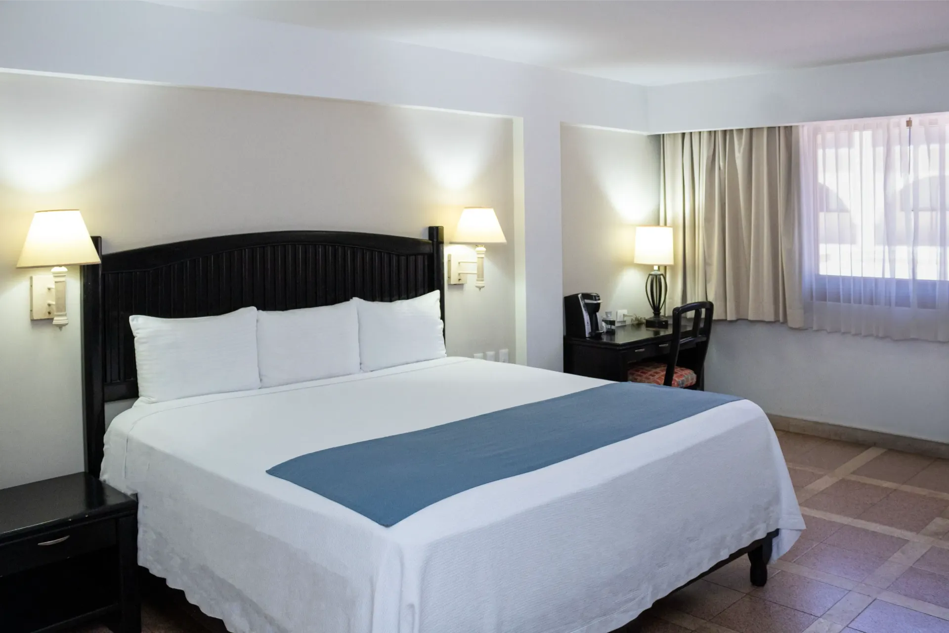 Habitación de hotel con cama, escritorio y ventana. Paredes blancas, camino de cama azul y baldosas color beige en el suelo.