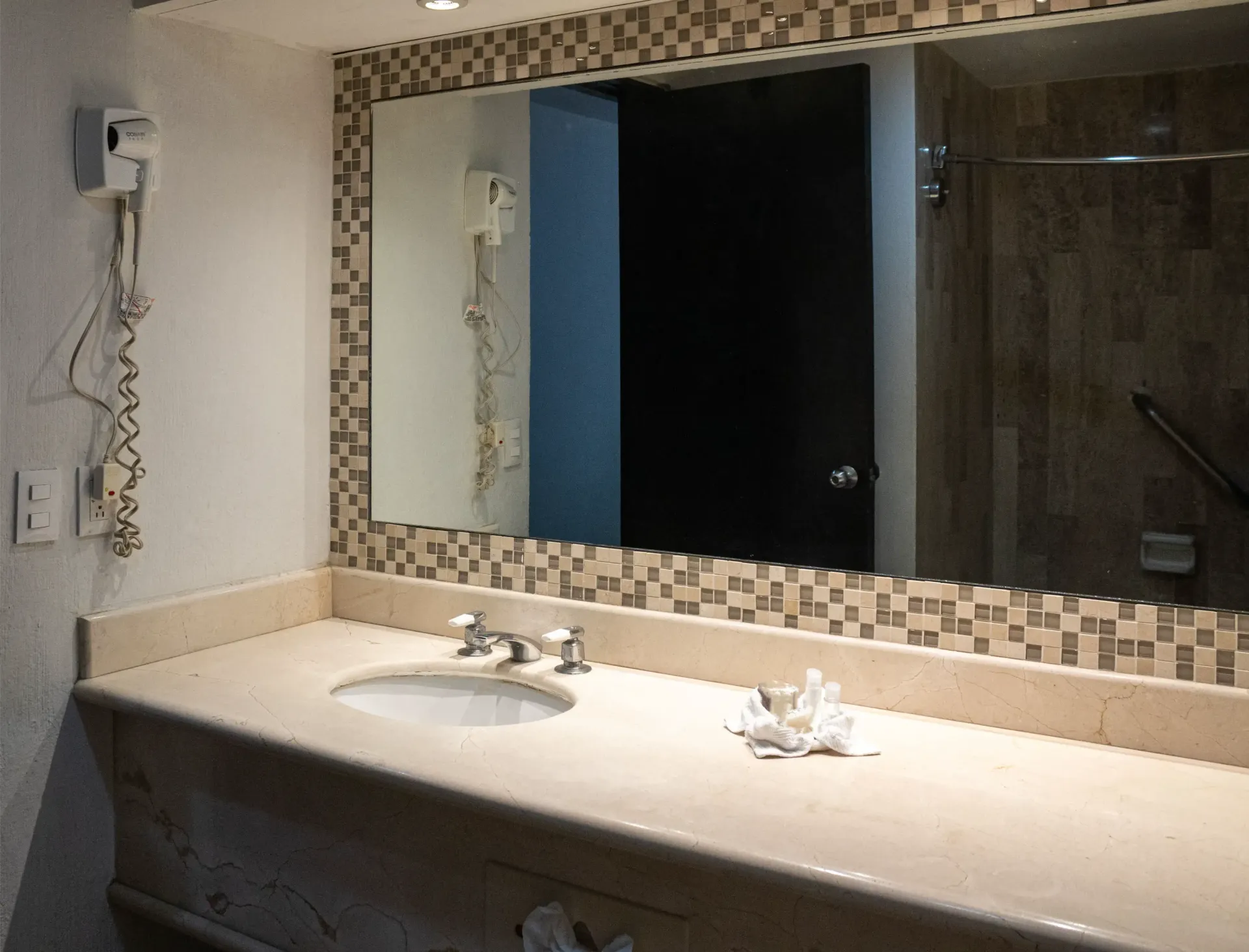 Tocador de baño con espejo grande, borde de azulejos de mosaico y lavabo integrado. Secador de pelo en la pared.
