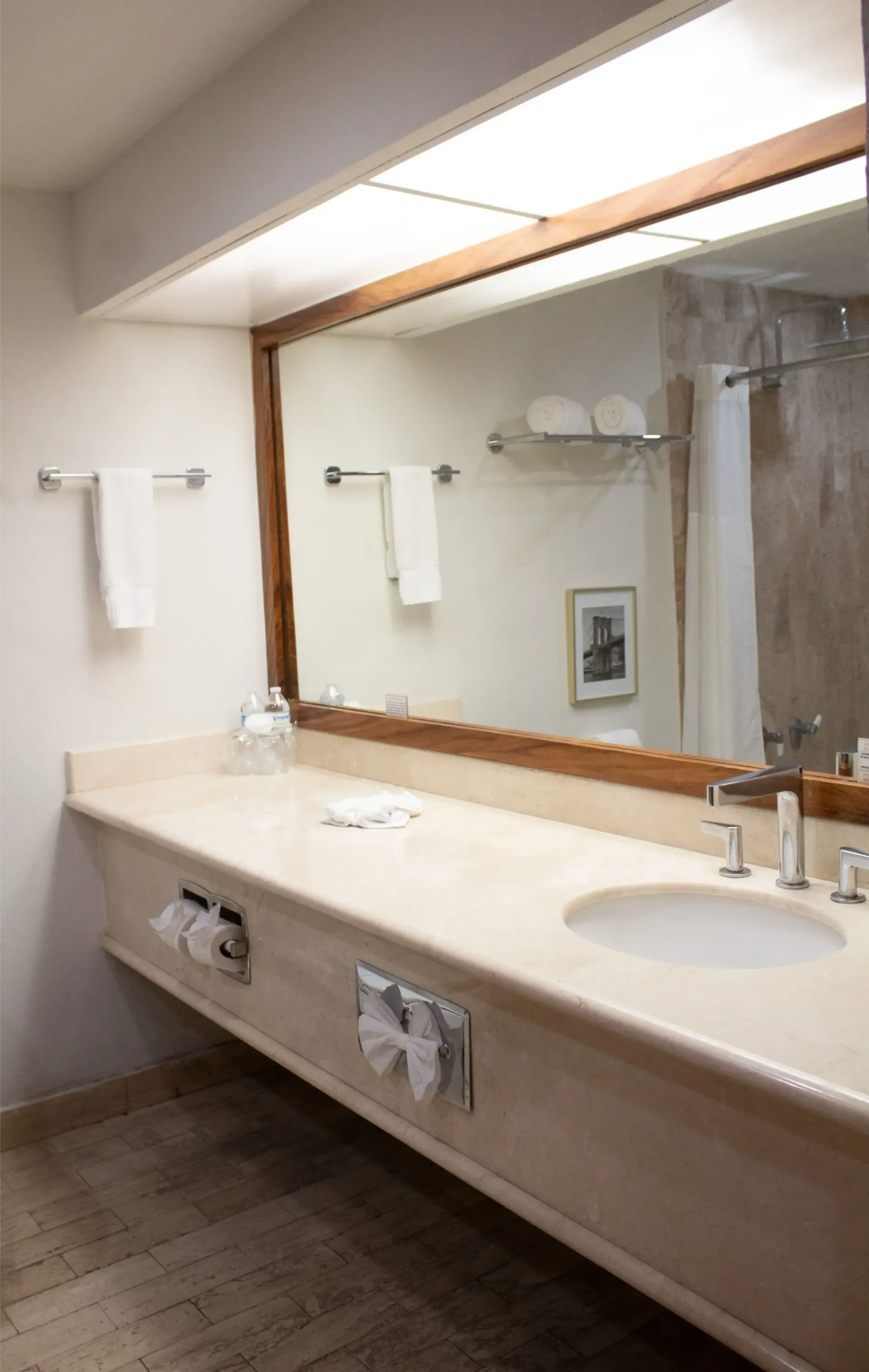 Baño con un gran espejo sobre una encimera de mármol. Se ven los toalleros, el lavabo y la ducha.