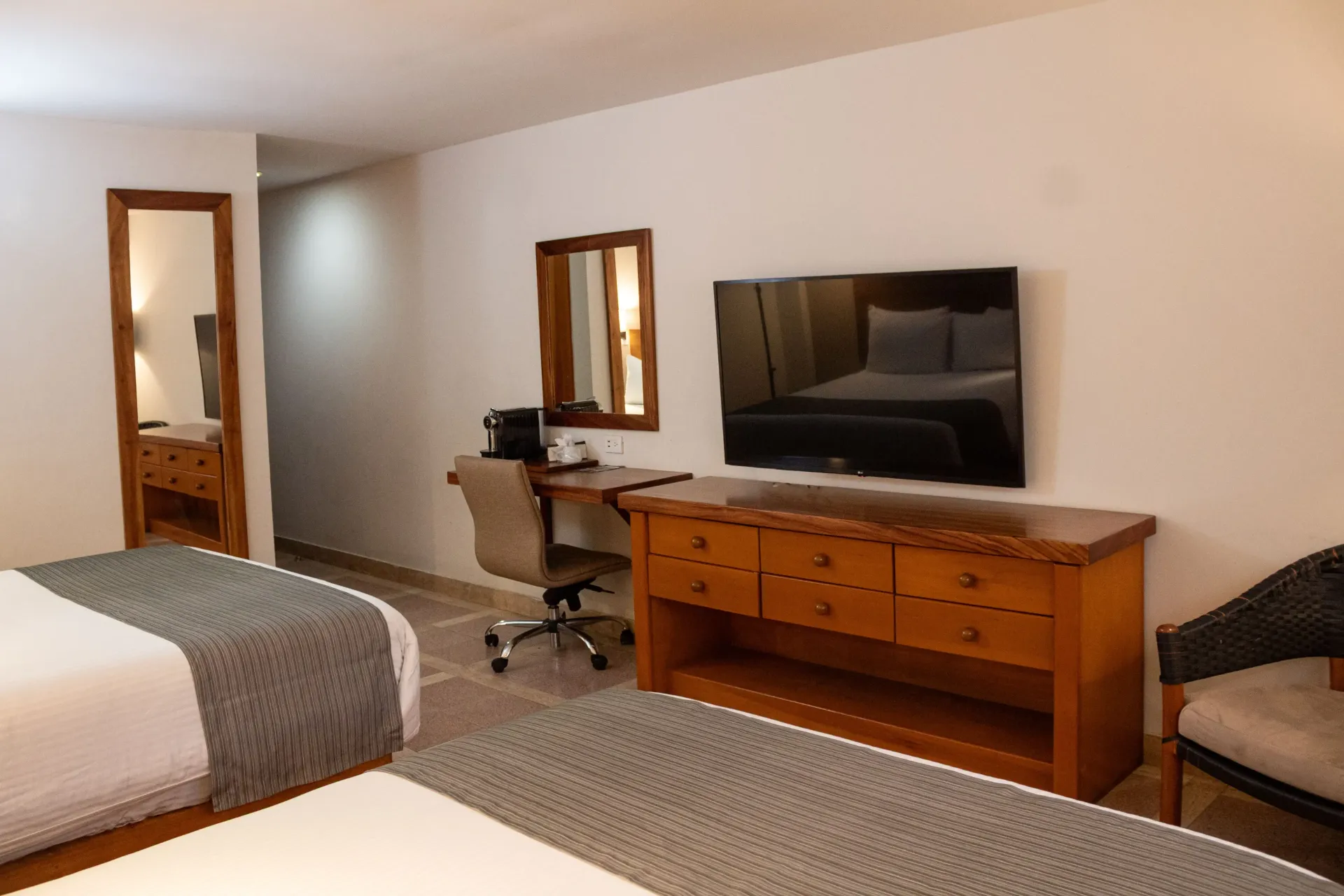Habitación de hotel con dos camas, TV, escritorio y espejo. Decoración en tonos tierra.