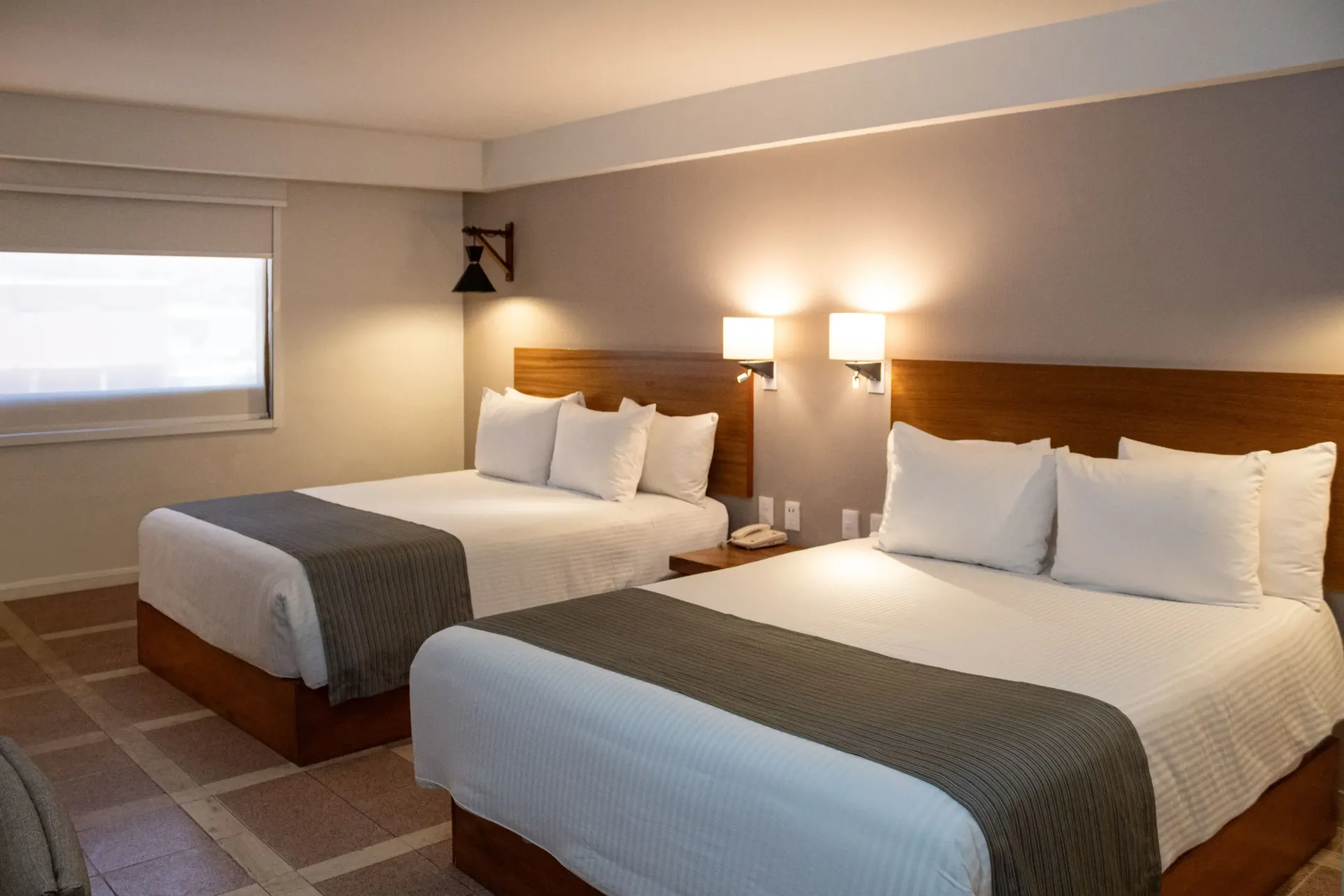Habitación de hotel con dos camas queen, ropa de cama blanca, cabeceros de madera y apliques de luz.