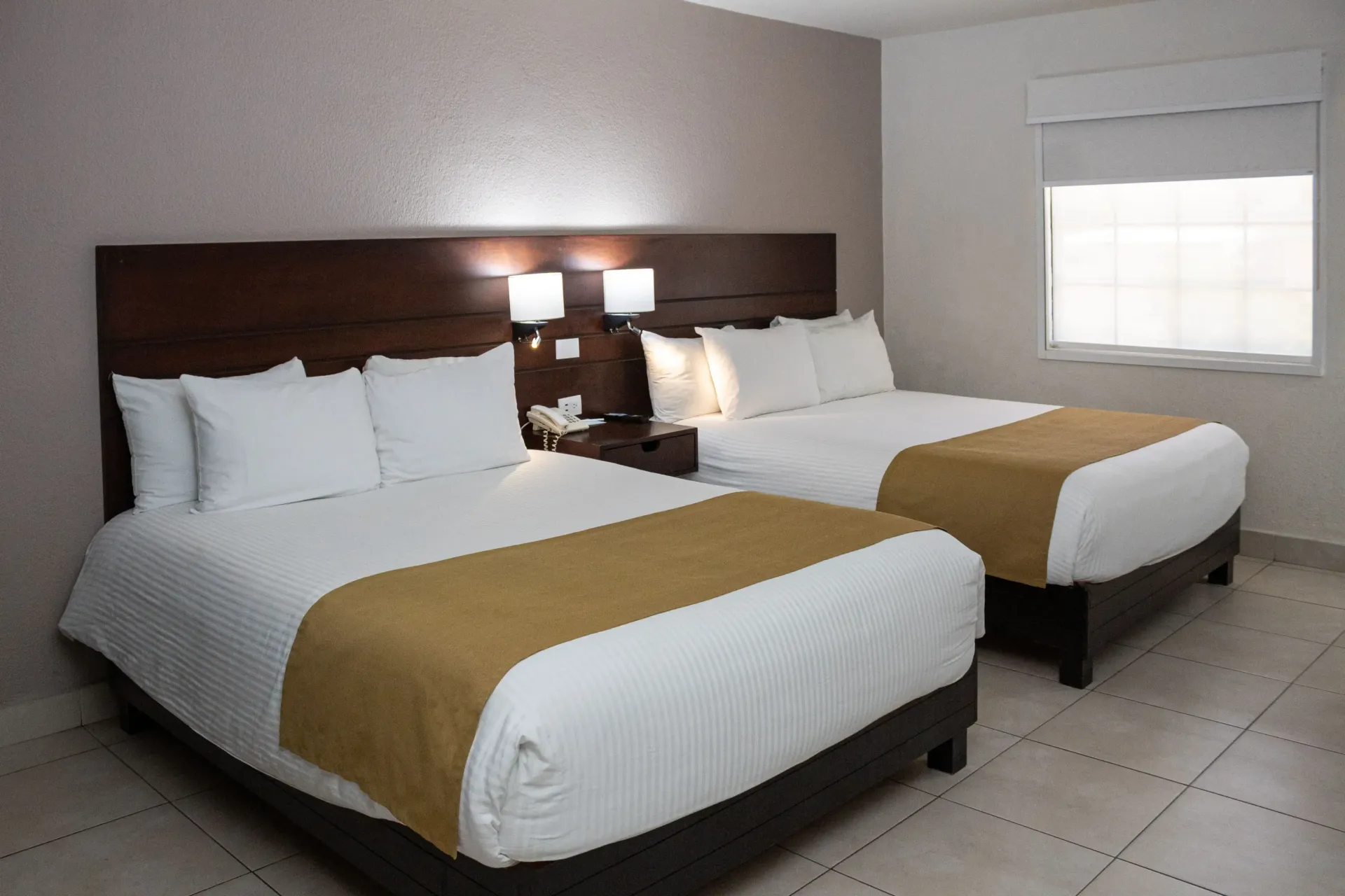 Habitación de hotel con dos camas dobles, cabecero marrón, sábanas blancas y caminos de cama color beige.