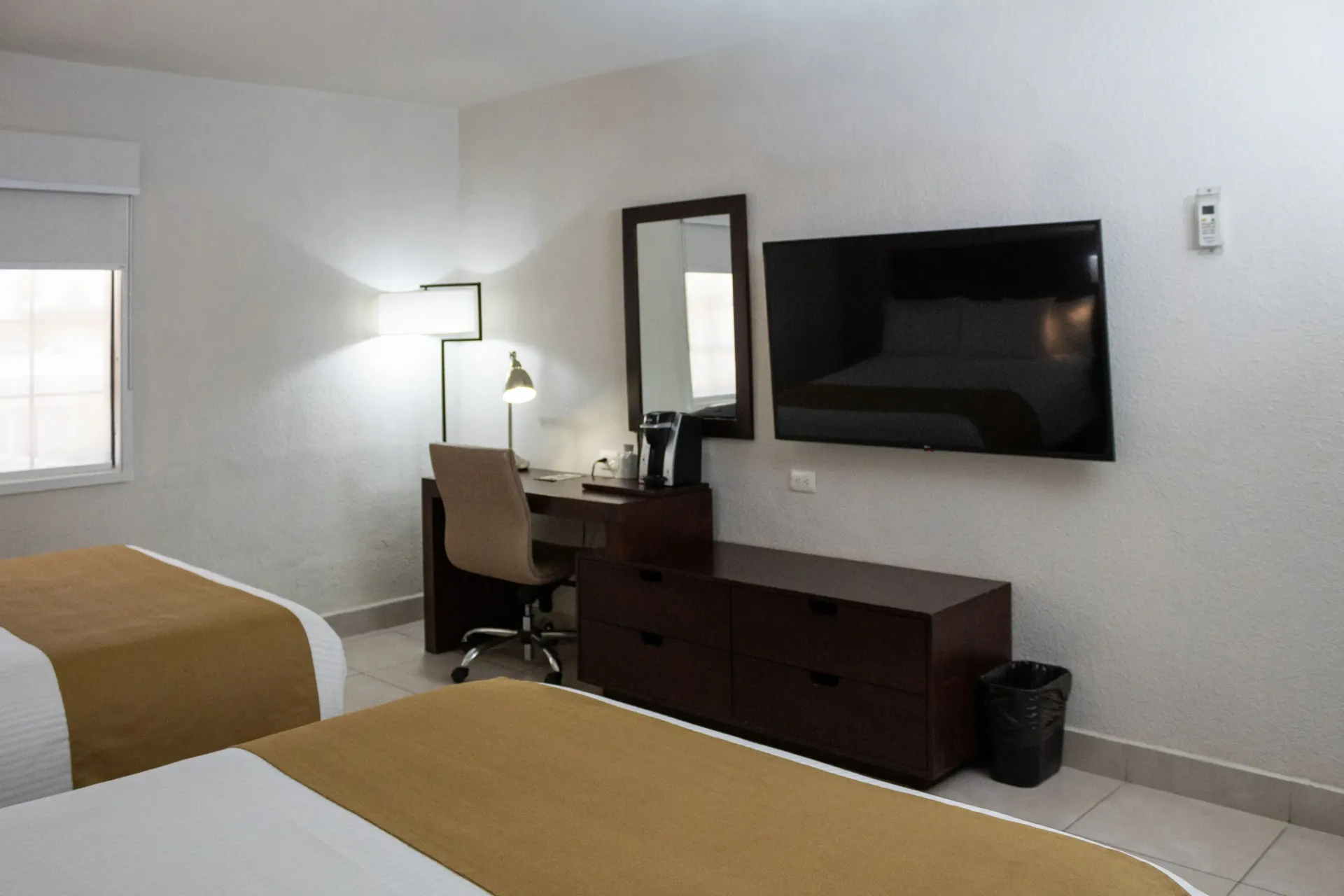 Habitación de hotel con dos camas, escritorio, espejo y televisor en una pared blanca.