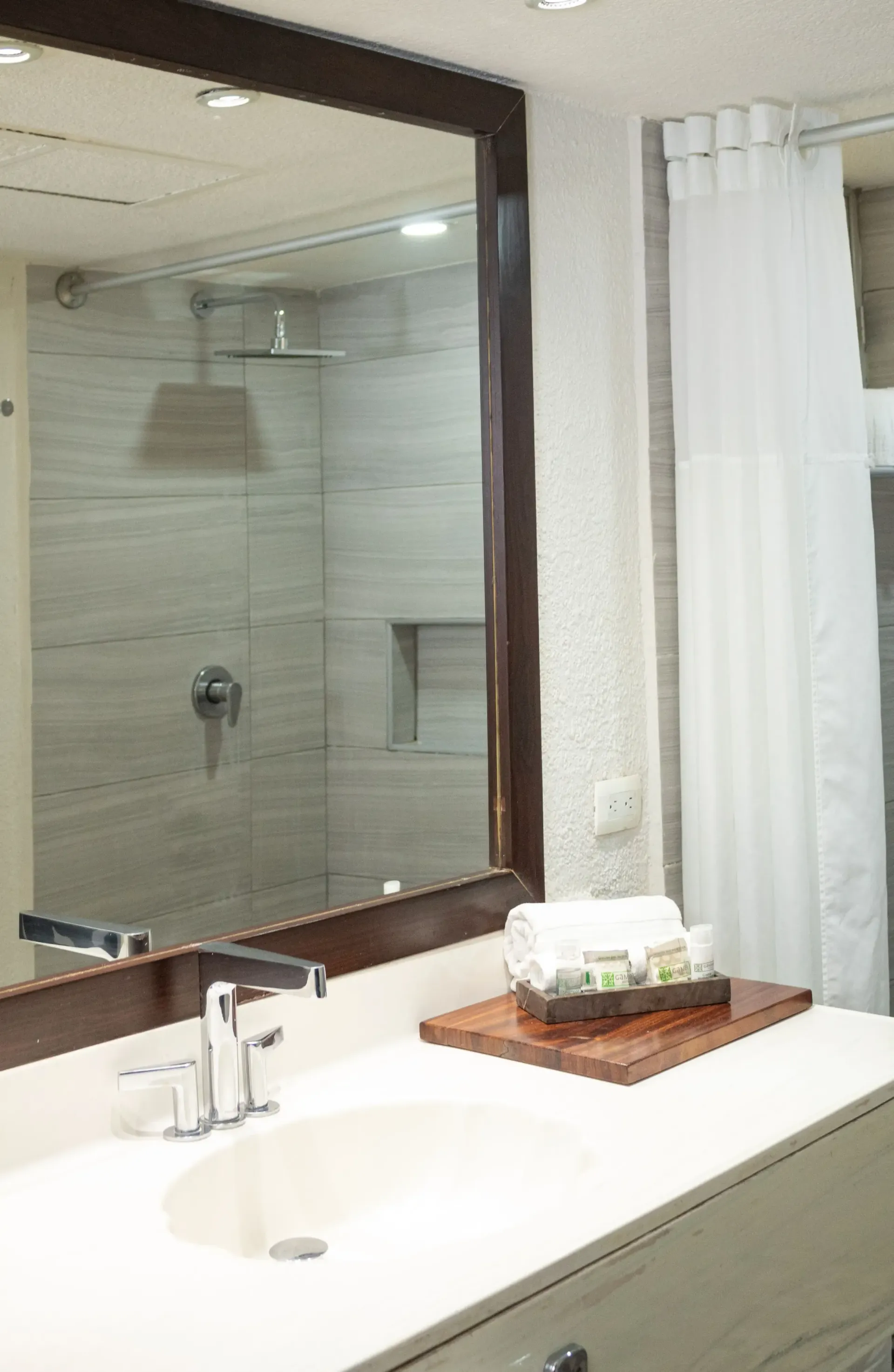 Baño con espejo grande, lavabo y ducha. Toallas blancas y artículos de aseo en una bandeja de madera.