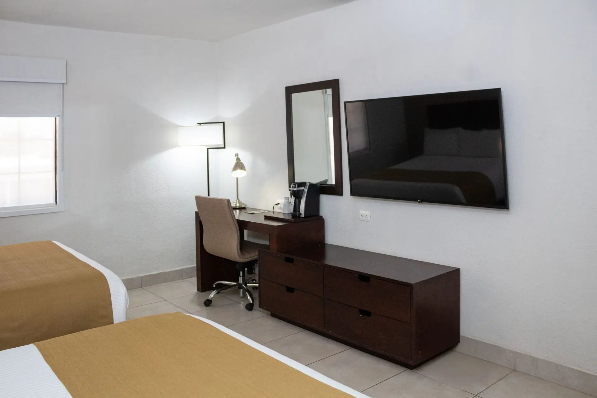 Habitación de hotel con dos camas, escritorio, televisor y un gran espejo en una pared blanca.
