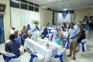 People seated at decorated banquet tables in a bright hall, with blue-and-white chair covers and tablecloths.