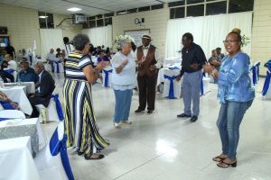 People standing and chatting in a bright hall with white tables and chairs, one dancing in a striped dress