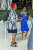 Two women dancing indoors, one in a blue dress and one in a zigzag top and red headwrap.