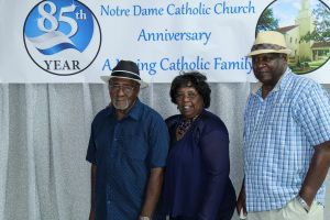 Three people pose in front of a Notre Dame Catholic Church 85th anniversary banner.