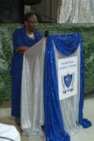 Woman speaking at a podium draped in blue and silver cloth, with a shield emblem on the front.