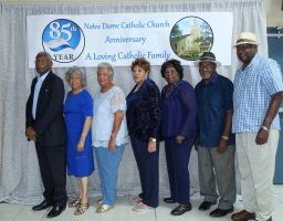 Seven people posing on a stage in front of a Notre Dame Catholic Church 85th anniversary banner.