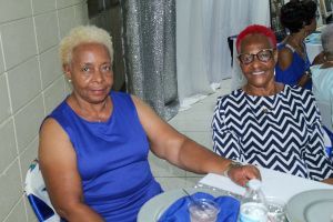 Two women seated at a table, smiling, with a blue cloth and water bottles in a tiled room