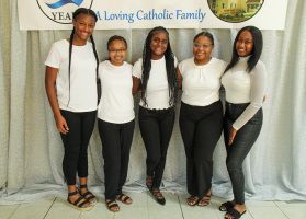 Five women standing in front of a Catholic family banner, wearing white tops and black pants.