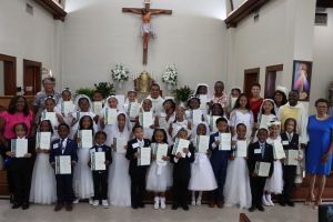 Group of children in white attire holding certificates in a church beneath a crucifix.