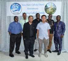 Six men posing in front of a Notre Dame Catholic Church anniversary banner