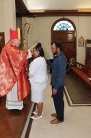 Three people stand indoors during a religious ceremony, facing a priest in red vestments.