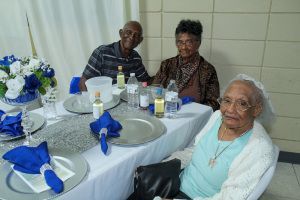 Three people seated at a decorated dining table with blue napkins and white plates in a banquet room