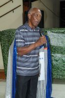Person holding a microphone and blue-and-white draped sash, standing indoors in front of greenery.