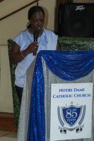 Woman speaking at a podium with a microphone beside a sign for Morris Dam Catholic Church.