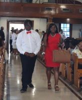 Two people walking down an aisle in a hall, one in a white shirt with a red bow tie and one in a red dress carrying a handbag
