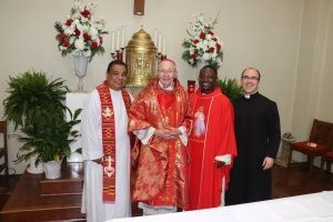 Four clergy in red and black vestments stand before an altar with flowers and a golden chair.