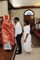 Church ceremony with priest in red vestments and two attendees standing beside him near the altar.