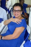 Woman in blue dress and black glasses seated indoors, smiling and touching her chest