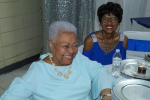 Two smiling women seated at a banquet table with silver dishes and a water bottle