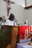 Person speaking at a church lectern beside a red-draped altar and crucifix.
