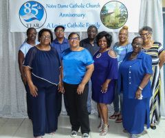 Group posing at Notre Dame Catholic Church anniversary banner, mostly dressed in blue.